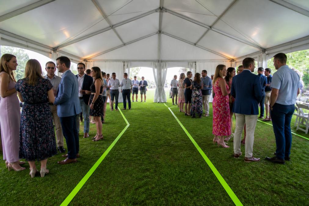 Outdoor tented event venue with clear exit routes marked by bright tape and signage, guests mingling under tent structure with proper spacing and emergency exits visible