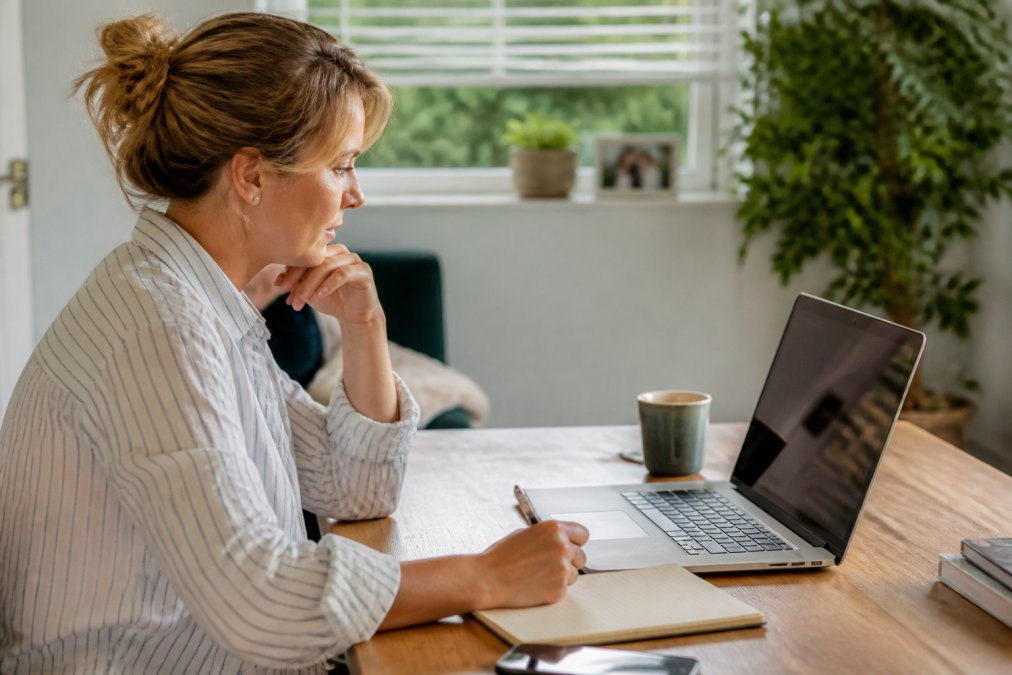 A local event organiser sitting at a desk reviewing ticketing platform options on a laptop, with a notepad and coffee nearby in a bright, naturally lit home office setting