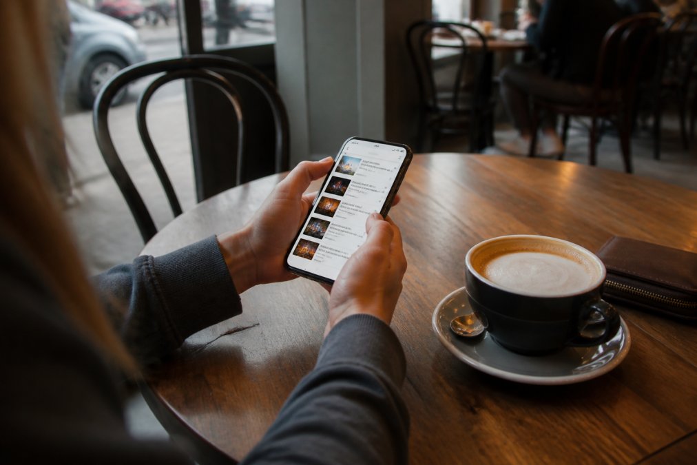 Person browsing events on a smartphone while sitting at a café table with a coffee, showing a relaxed event discovery experience with visible event listings on screen