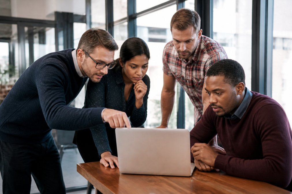 Team of database engineers collaborating around a laptop displaying colorful execution plan trees with cost metrics, query statistics, and performance graphs in a modern office with natural window lighting