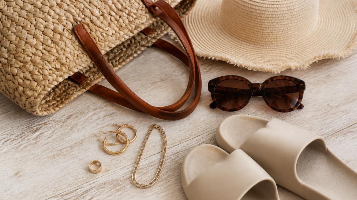 A flat lay on a light timber surface showing a woven beach tote, oversized sunglasses, a wide-brimmed hat, simple gold jewellery and a pair of comfortable slides, styled together as a coastal accessory set