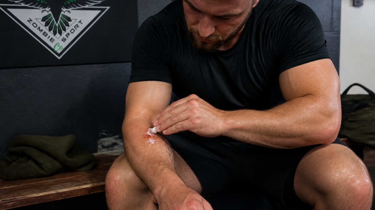Combat athlete applying Regenerate RX balm directly to a fresh scrape on their forearm immediately after training, showing the product in active use with proper wound contact