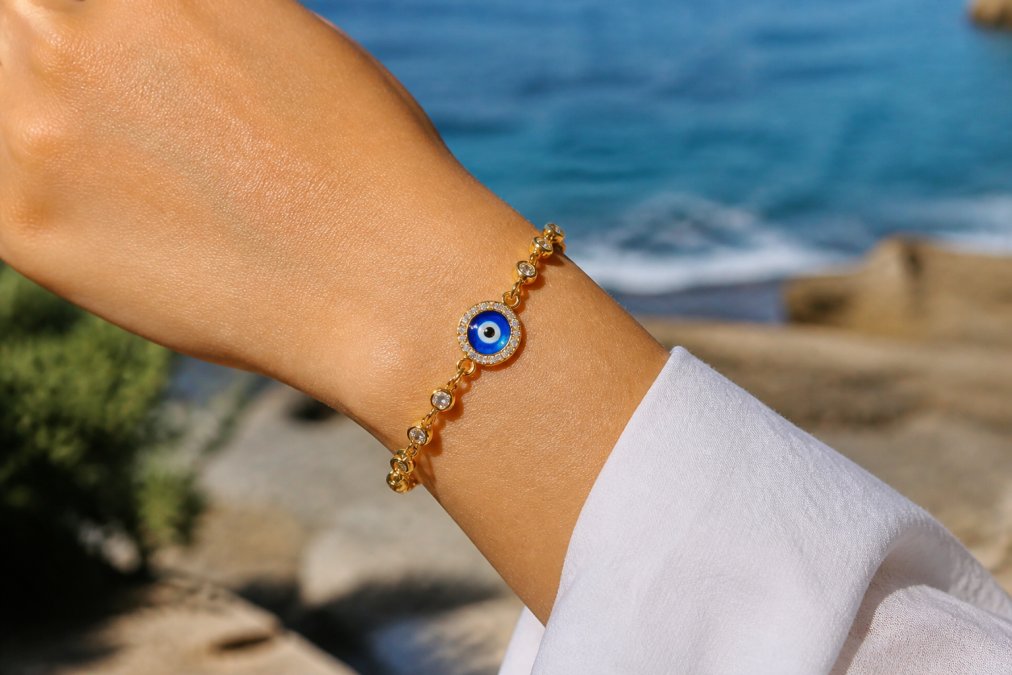 Close-up of a woman's wrist wearing a delicate gold-plated evil eye bracelet with vibrant blue enamel and cubic zirconia stones, showing the intricate craftsmanship and shimmer in natural daylight