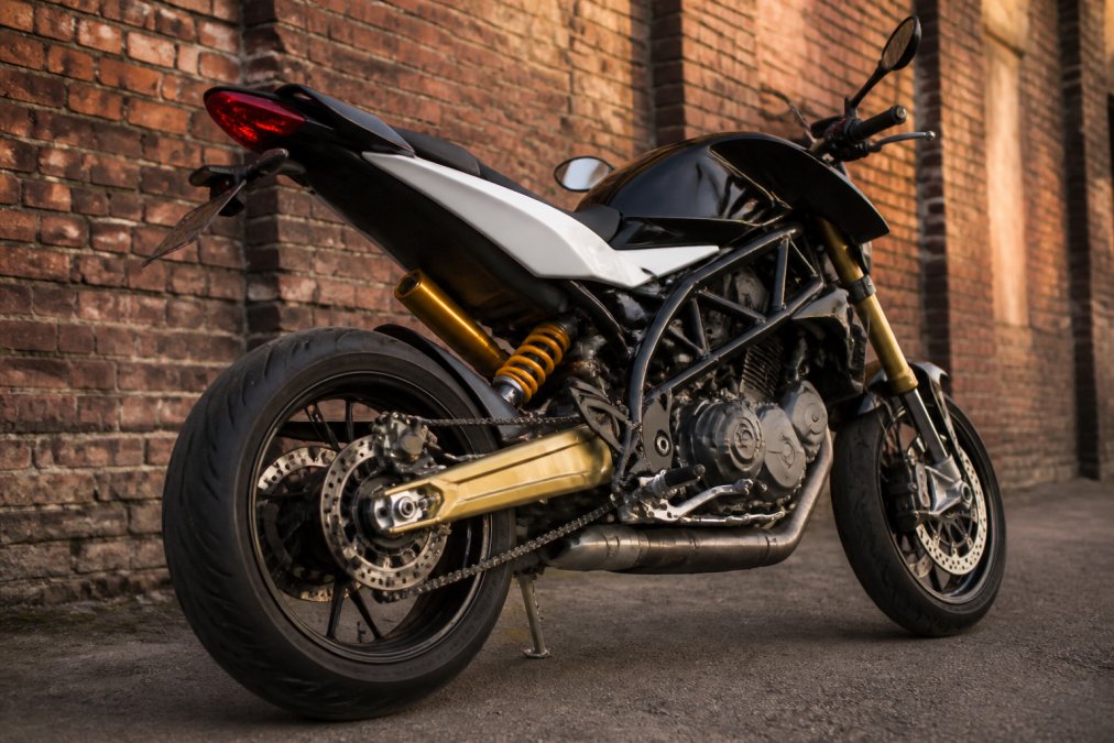 Close-up van een Husqvarna Nuda 900R met zichtbare gouden Öhlins achtervork en verende elementen, geparkeerd voor een industriële bakstenen muur bij warm avondlicht