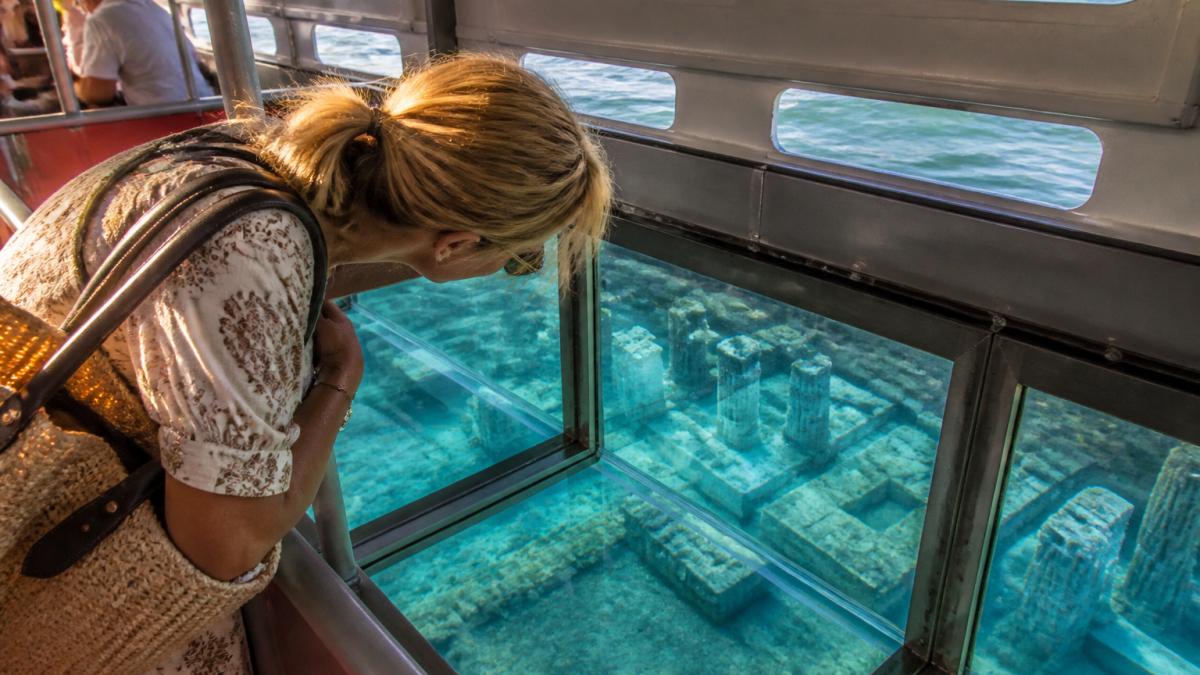 Tourists view submerged Roman ruins in Baia with exclusive access historical sites in Italy via glass-bottom boat.