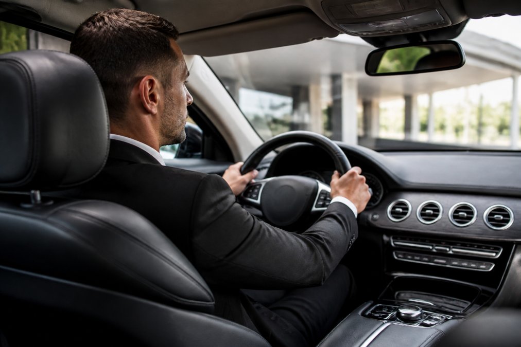 Luxury sedan interior showing a well-dressed professional driver at the wheel with hands positioned correctly on the steering wheel, clean leather upholstery, modern cabin with subtle ambient lighting, and professional interior environment