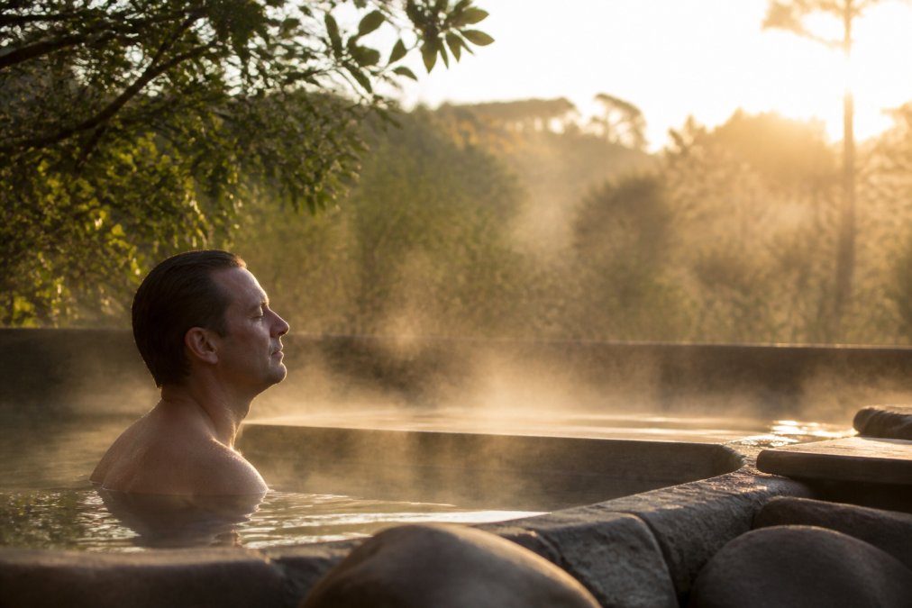 Executive in peaceful retreat setting, sitting by natural thermal waters at dawn, eyes closed in meditation, morning mist rising, soft golden light filtering through trees