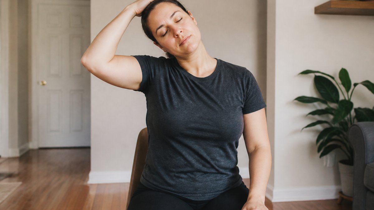 Person seated upright performing gentle neck stretch with hand supporting head tilt toward shoulder, good posture, natural indoor lighting
