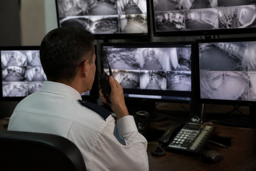 A uniformed security officer in a professional control room at night, monitoring multiple CCTV screens showing various property feeds, with alert indicators visible on two of the screens and a radio handset on the desk