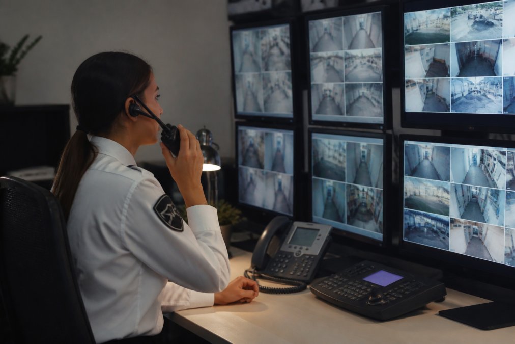 Security control room with multiple monitoring screens displaying alarm status and property feeds, dispatch operator at workstation coordinating response with radio headset and alert systems visible