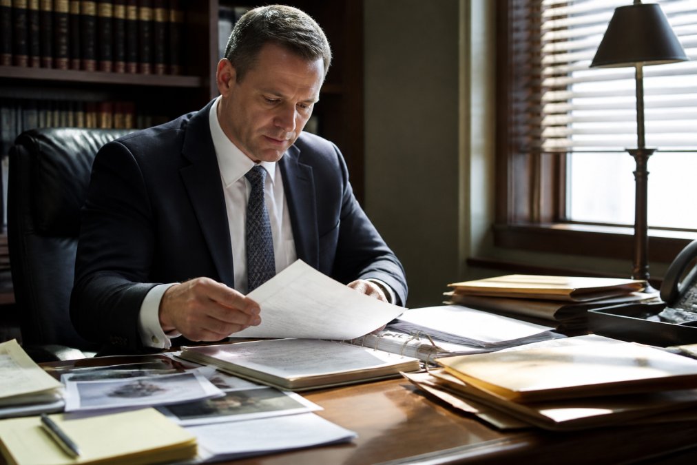 Defense attorney in professional dark suit reviewing case files and evidence documents at wooden desk in law office, surrounded by legal folders and case materials, natural window lighting