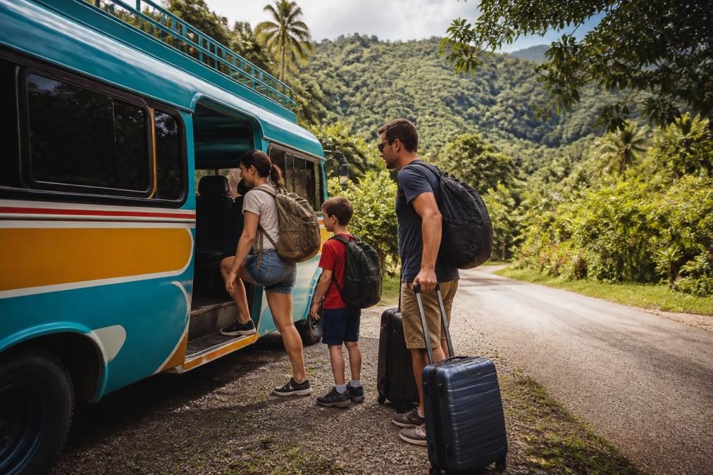 Family boarding a colorful local shuttle bus with luggage, surrounded by lush green landscape and tropical vegetation