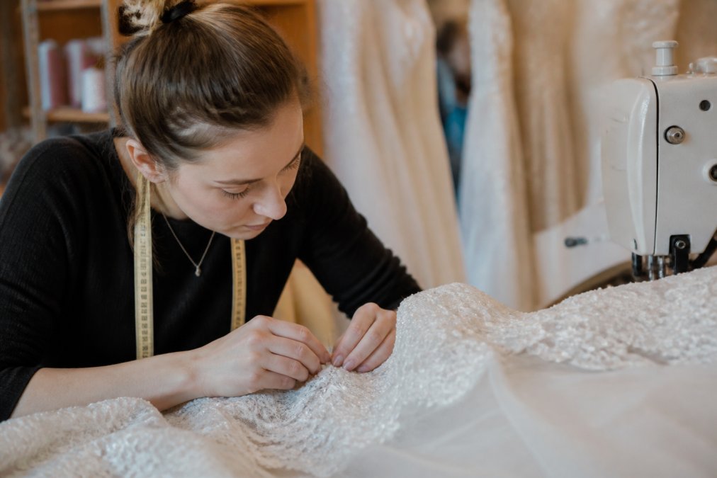 Professional bridal seamstress working at a sewing machine, carefully adjusting a white wedding gown with precision, pinning delicate lace details while focused on the intricate beadwork