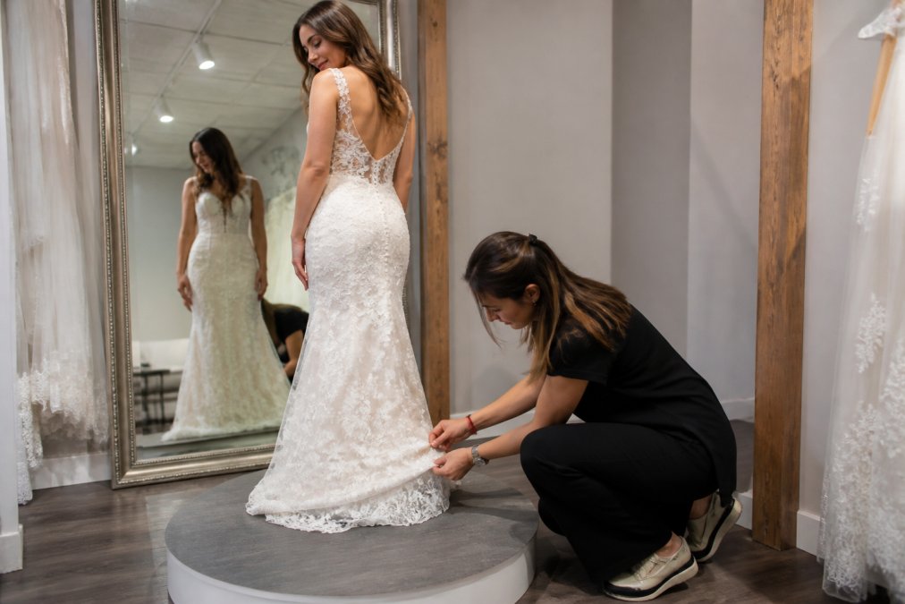 Bride standing on a fitting platform in front of a large mirror while a professional seamstress measures and carefully pins the hem of a white wedding dress, adjusting the length with precision