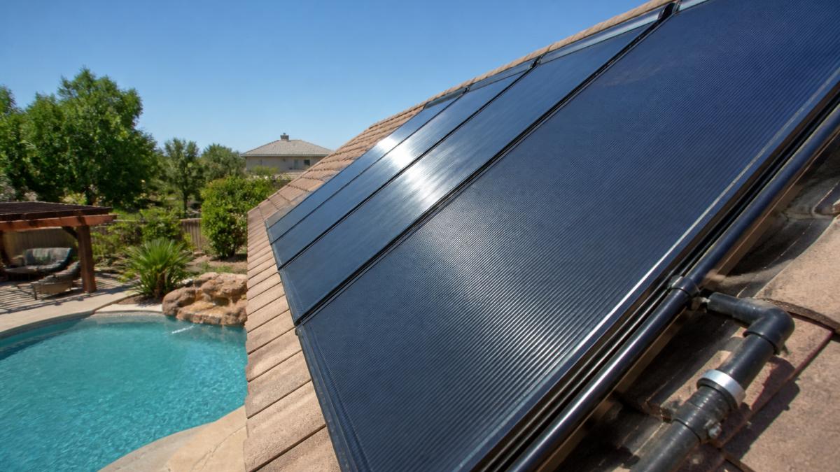 Close-up of residential solar pool heater panels mounted on roof with pool visible below, showing installation in sunny backyard setting with clear blue sky