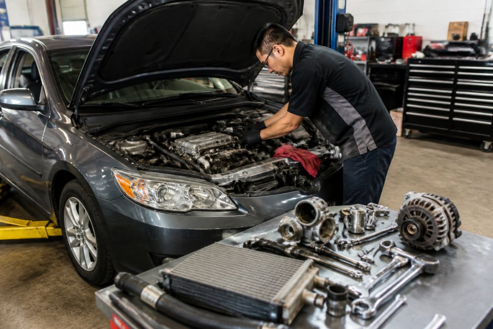 Professional technician in a well-lit repair bay examining the engine of a high-mileage Japanese sedan, with diagnostic equipment, wrenches, and replacement parts visible on the workbench