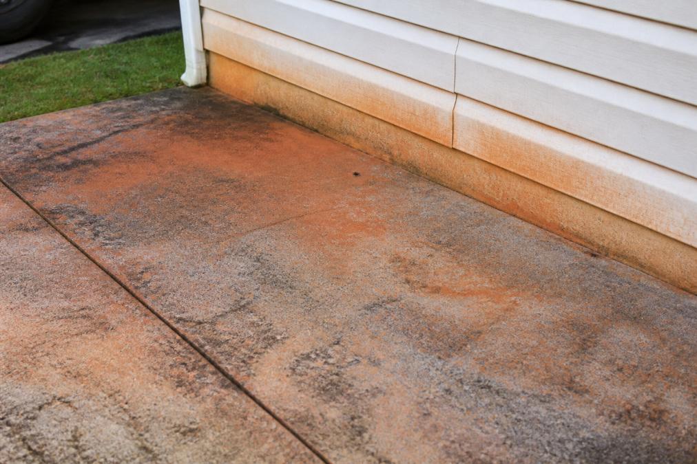 Close-up of red clay stains on residential driveway and siding showing stubborn discoloration typical of regional soil conditions, with weathered concrete surface
