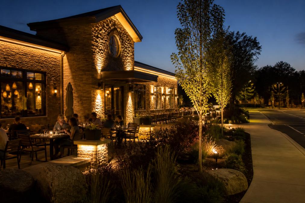 A restaurant patio at dusk with warm exterior lighting illuminating the building facade, tables, and landscaping, with customers dining and the business sign glowing softly