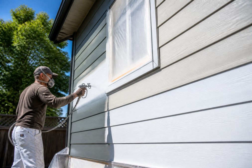 Professional painter applying high-quality exterior paint to residential home siding using spray equipment on a bright sunny day with clear blue sky