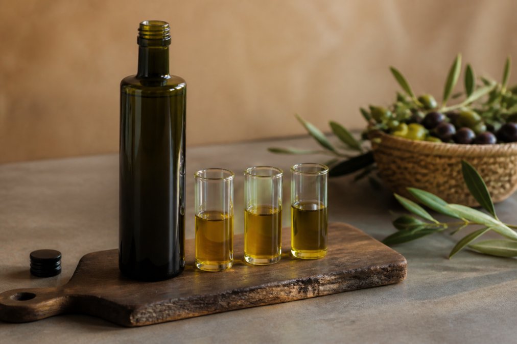 Een geopende fles Forelia "2 Roots" extra vierge olijfolie naast drie kleine proefglaasjes met olie van verschillende variëteiten, op een donker houten plankje