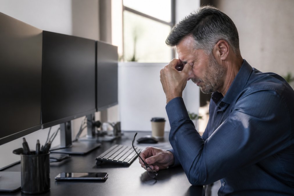 Middle-aged professional at desk with multiple monitors, rubbing eyes in visible fatigue and discomfort, natural office lighting showing strain on face