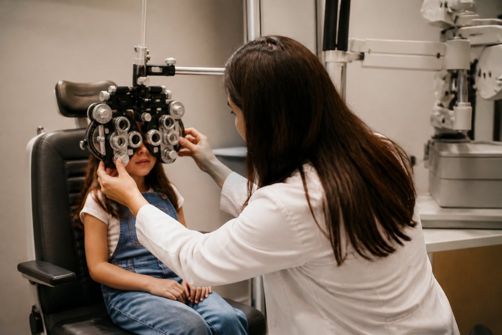 A young child sitting in an optometry chair while an optometrist uses a phoropter to assess their vision, with warm office lighting and the child appearing calm and engaged