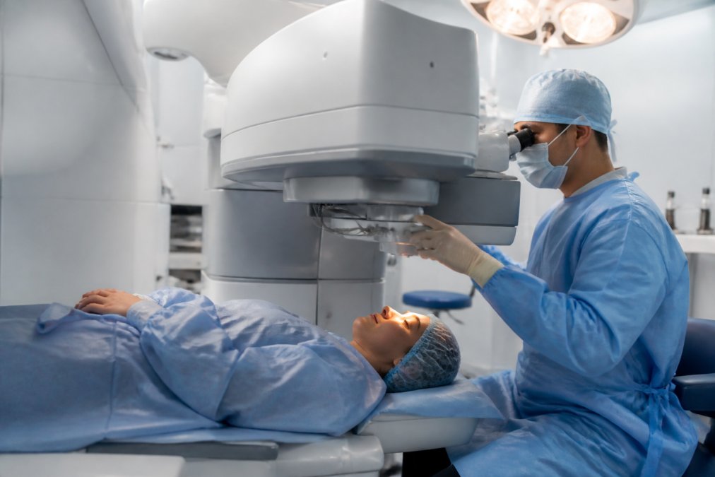 A modern ophthalmic surgical suite with an advanced laser system positioned over a reclined patient bed, a surgeon in sterile attire adjusting equipment under bright operating lights