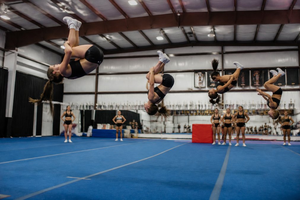 Professional cheer gym with athletes performing tumbling passes on a high-performance spring floor, showing confident execution of aerial skills with proper impact absorption visible in body positioning