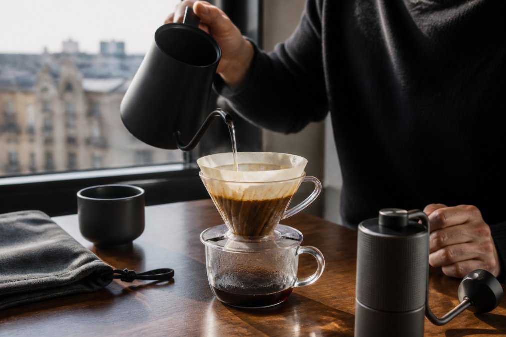 Nærbillede af hænder, der forsigtigt hælder varmt vand over kaffegrums i et håndbrygningsudstyr på et hotelværelsebord, med stille morgenlys gennem vinduet