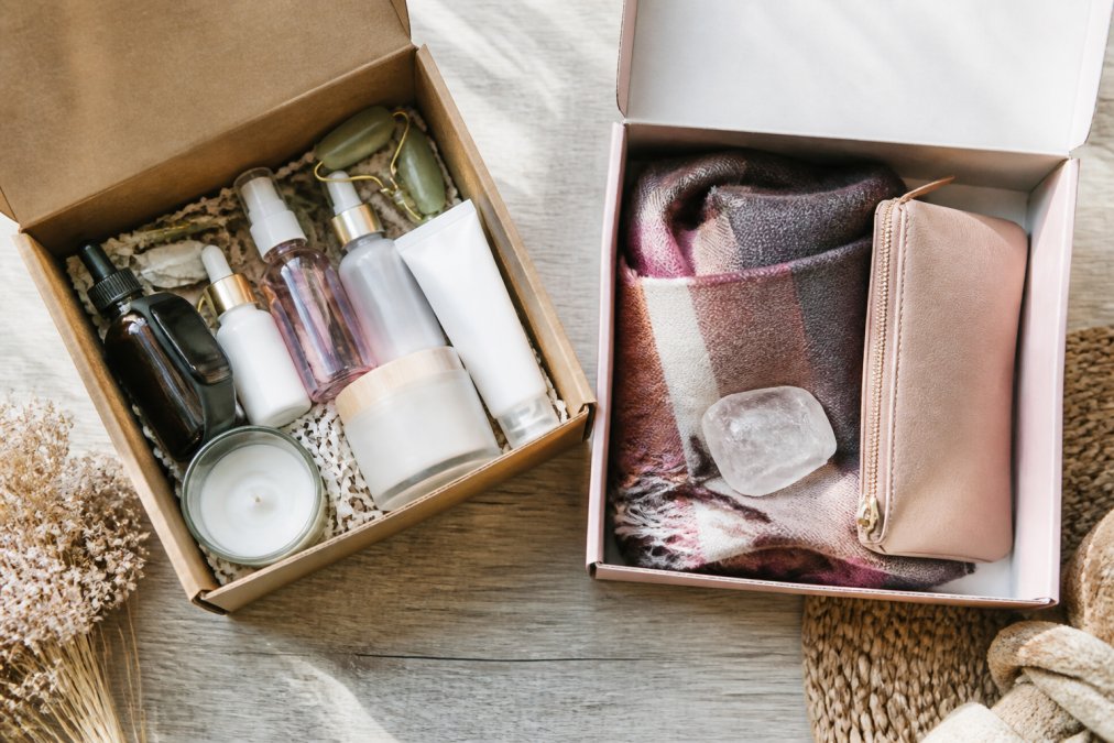 Overhead flat lay of two open subscription boxes on a light wood table: one filled with skincare bottles, a fitness band, and a small candle; the other with a folded scarf, a crystal, and a cosmetic pouch. Soft natural light, styled for a lifestyle blog
