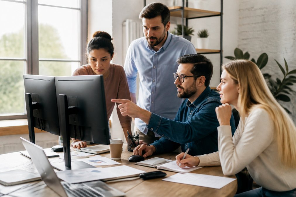 Startup founder reviewing performance dashboards and analytics reports on multiple monitors with team members collaborating around the desk in a modern office with natural window lighting