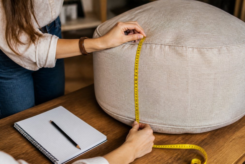 Close-up of hands holding a measuring tape against a beige pouf, with a notebook and pencil nearby on a wooden work surface