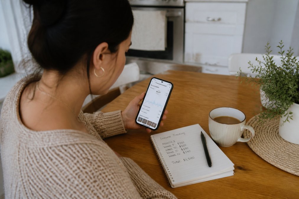 A woman sitting at a kitchen table with a smartphone showing Instagram analytics, a notebook with handwritten income tracking, and a coffee mug nearby