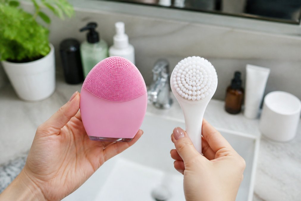 Close-up of a person holding a silicone facial cleansing brush in one hand and a manual bristle brush in the other, with a bathroom sink and skincare products in the background