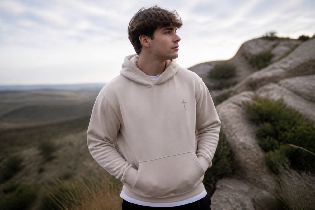 A young adult wearing a minimalist faith-based hoodie with a small cross emblem, standing outdoors in natural light, casual and modern styling