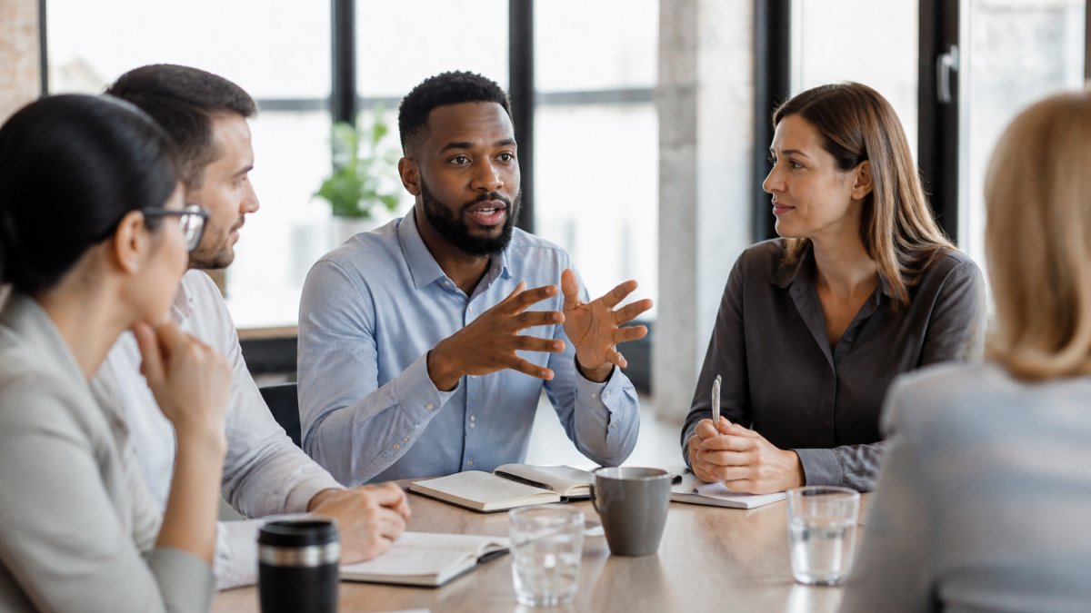 Diverse group of professionals sitting around a conference table engaged in discussion, with one person gesturing while speaking, natural office lighting streaming through windows, collaborative atmosphere