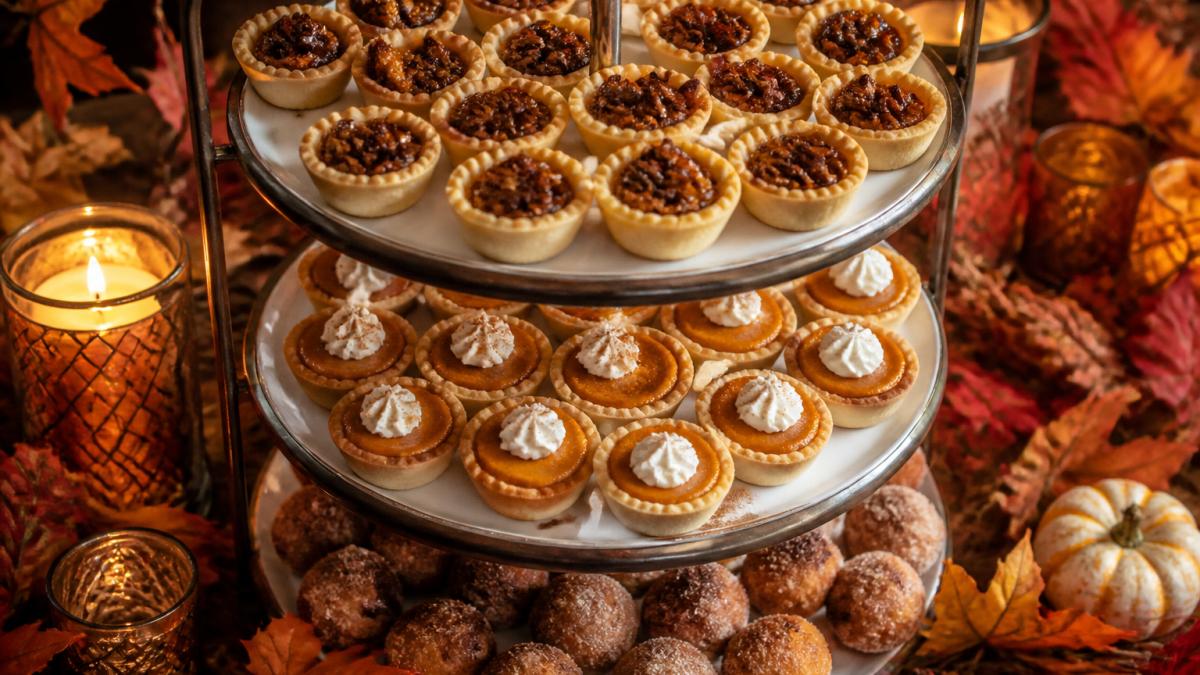 Overhead shot of an elegant fall dessert display featuring mini pecan pies, pumpkin tartlets, and apple cider donut holes arranged on a tiered stand with autumn foliage, candles, and warm amber lighting