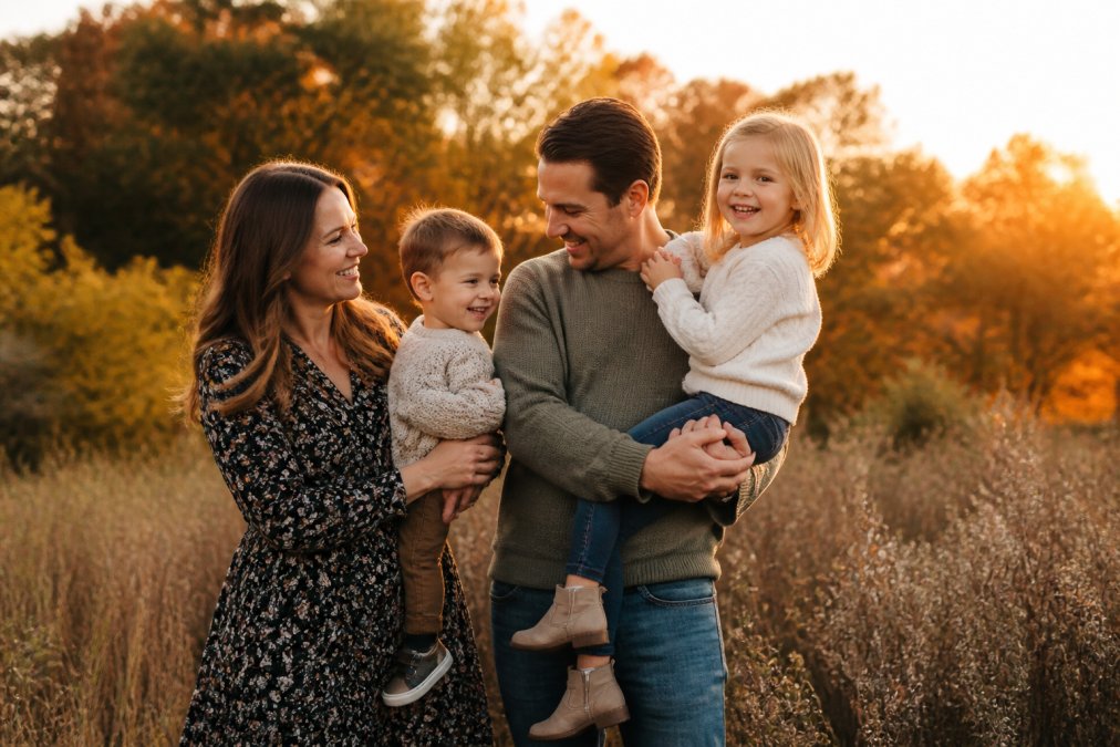 A relaxed family of four laughing together outdoors during golden hour, with autumn foliage in soft focus behind them, capturing natural movement and genuine connection
