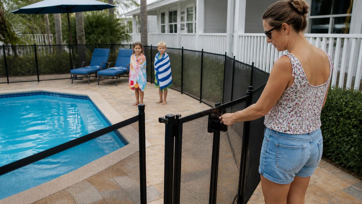 Parent testing a self-latching gate for private pool safety while children wait on the deck with beach towels