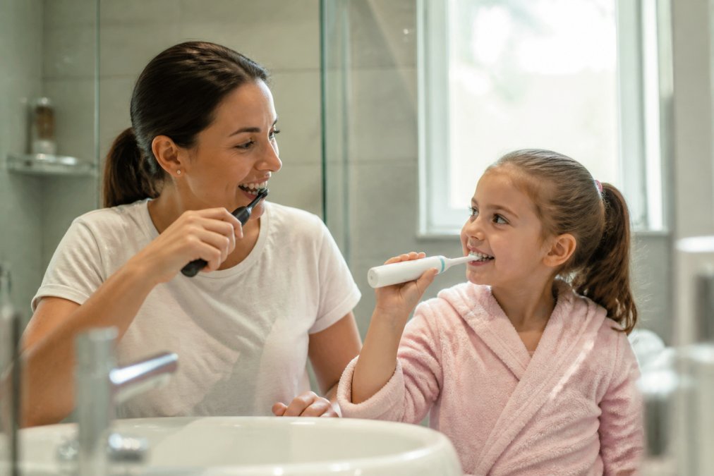A parent and child brushing their teeth together in a bright, modern bathroom, each using a different type of electric toothbrush, with natural morning light streaming through the window