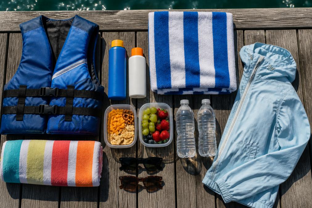 Overhead view of boat tour essentials laid out on a dock: sunscreen bottles, life jackets, towels, snacks in containers, water bottles, sunglasses, and a light jacket arranged neatly on weathered wood