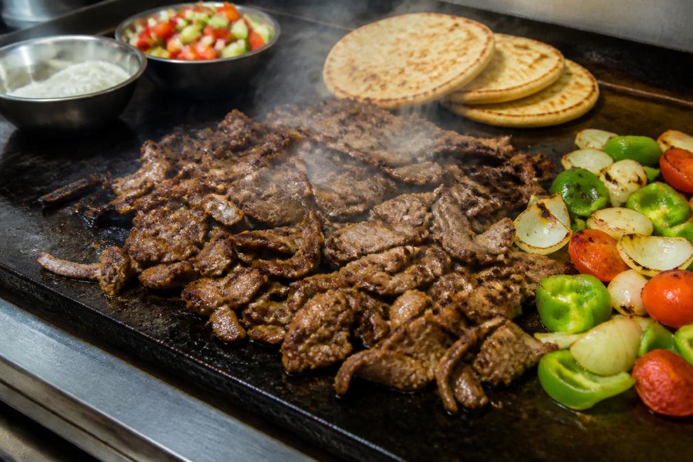 Vibrant overhead shot of freshly grilled gyro meat sizzling on a grill with colorful vegetables, pita bread, and tzatziki sauce arranged nearby, steam rising from the hot surface, professional kitchen lighting