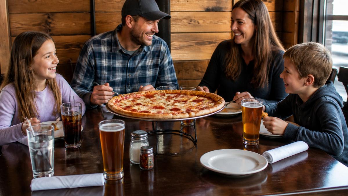 Family of four enjoying fresh pizza and drinks at a casual restaurant table with children smiling and engaged, warm lighting and relaxed dining atmosphere with wood-accented walls