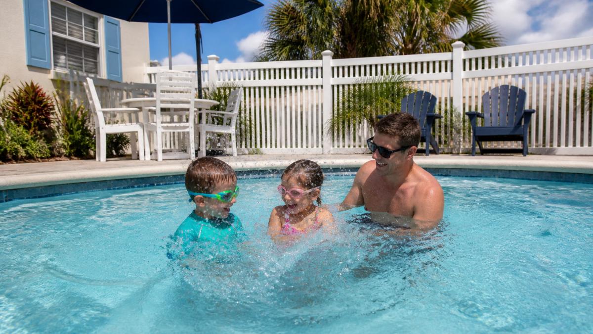 A family with two young children splashing in a private pool at a beach cottage, with a fenced patio and outdoor seating visible in the background