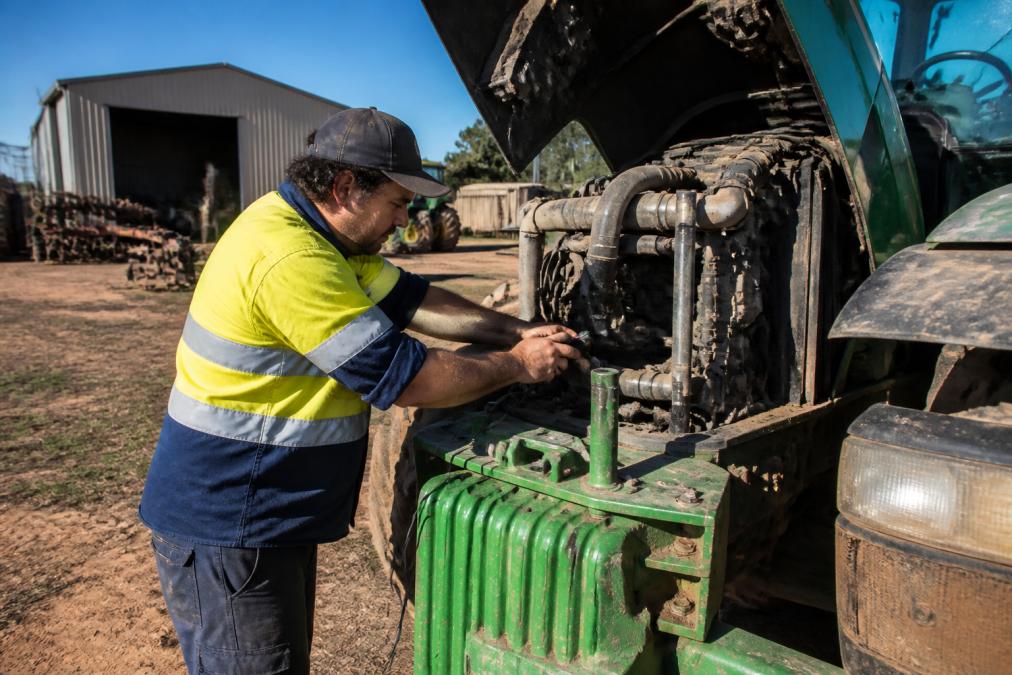 Mechanic in high-visibility vest performing diagnostic scan on a diesel tractor engine in a paddock, with farm equipment and storage sheds visible in the background under natural daylight