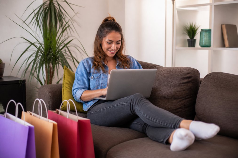 Woman browsing trendy dresses and jumpsuits on a laptop while sitting on a couch with shopping bags nearby, showing online boutique shopping experience