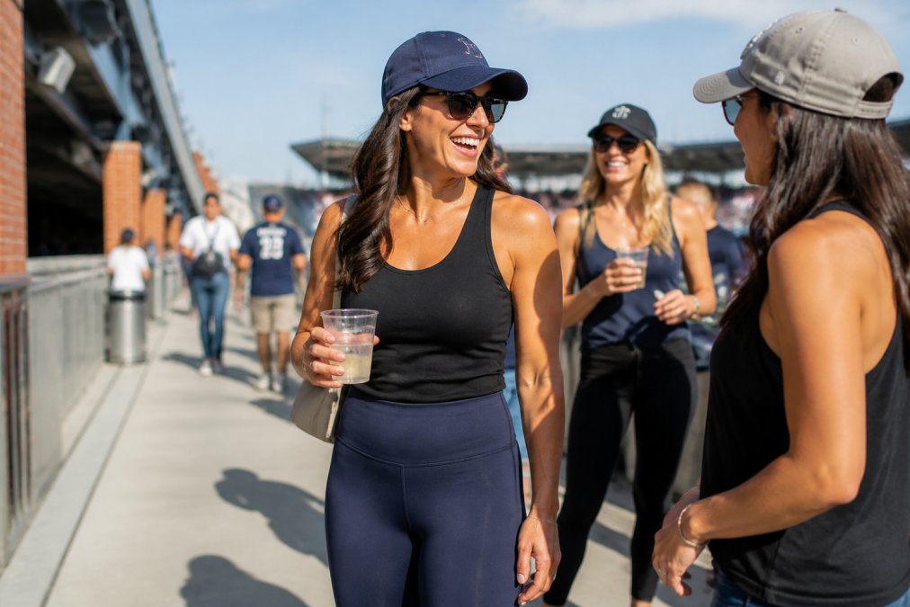 Woman in high-waisted leggings and a racerback tank standing in a sunlit stadium concourse with friends.