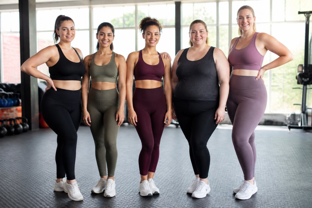 Diverse group of women wearing different styles of fashionable activewear that complement their individual body shapes, photographed in a bright, modern gym studio with natural lighting, showing confident postures and various silhouettes