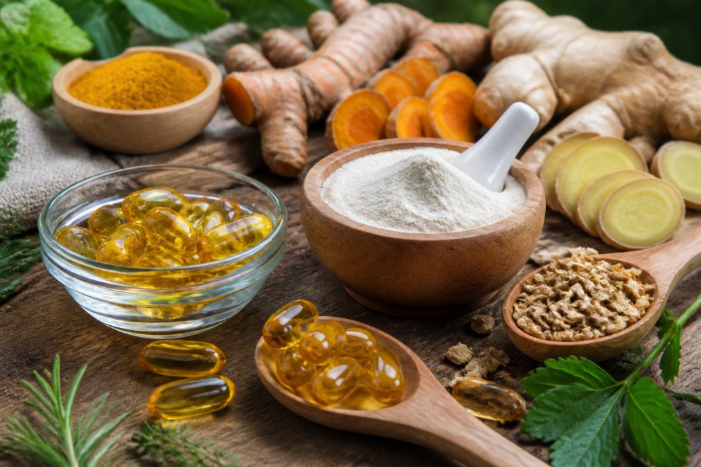 Close-up of natural supplement ingredients including turmeric root, ginger slices, omega-3 capsules, and collagen powder arranged on a wooden surface with warm natural lighting and fresh herbs