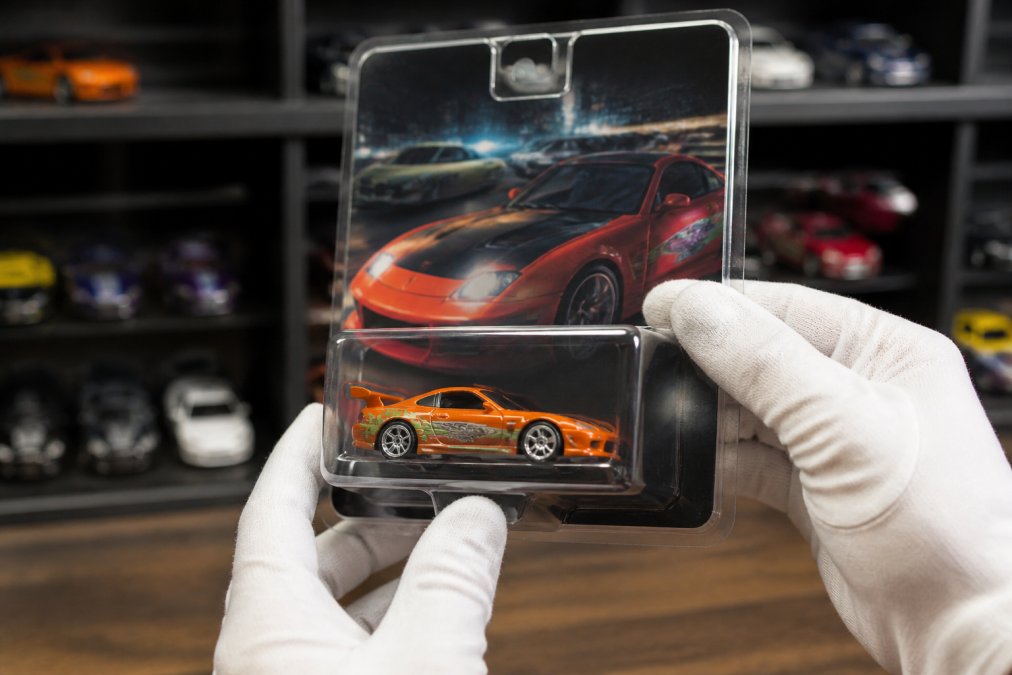 Close-up of hands in cotton gloves holding a factory-sealed Hot Wheels Fast and Furious car, the clear plastic clamshell reflecting soft light, with vibrant tampo graphics and intact cardback visible